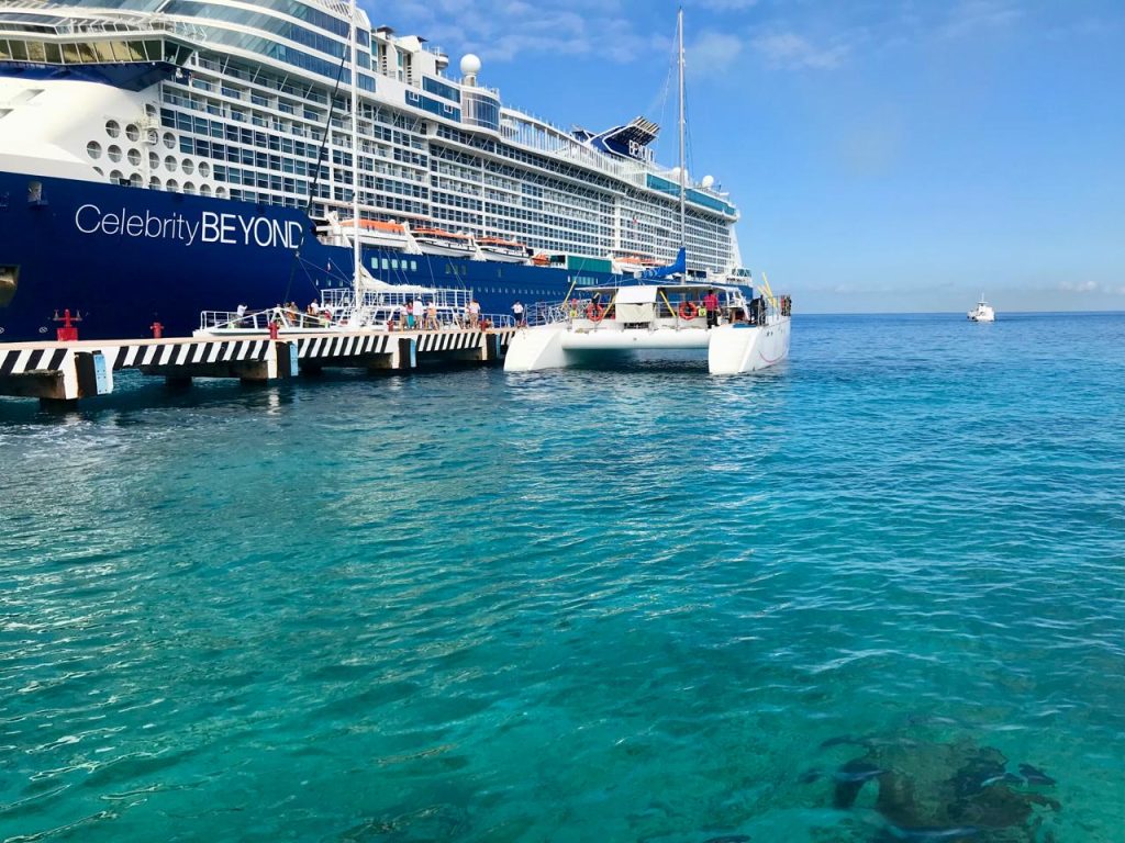 Celebrity Beyond cruise ship docked at a pier, with a catamaran preparing for departure in turquoise waters, showcasing a vibrant tropical setting.